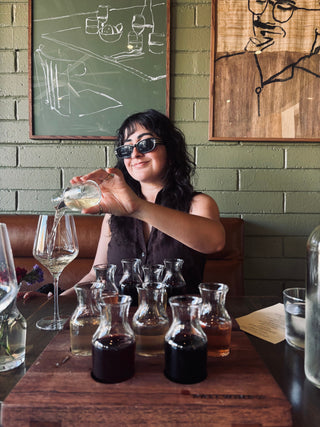Woman pouring a drink at a bar with various bottles and glasses on the counter.