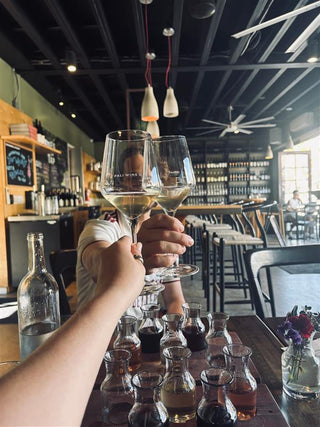 Two people clinking wine glasses in a bar setting with bottles and glasses on the table.